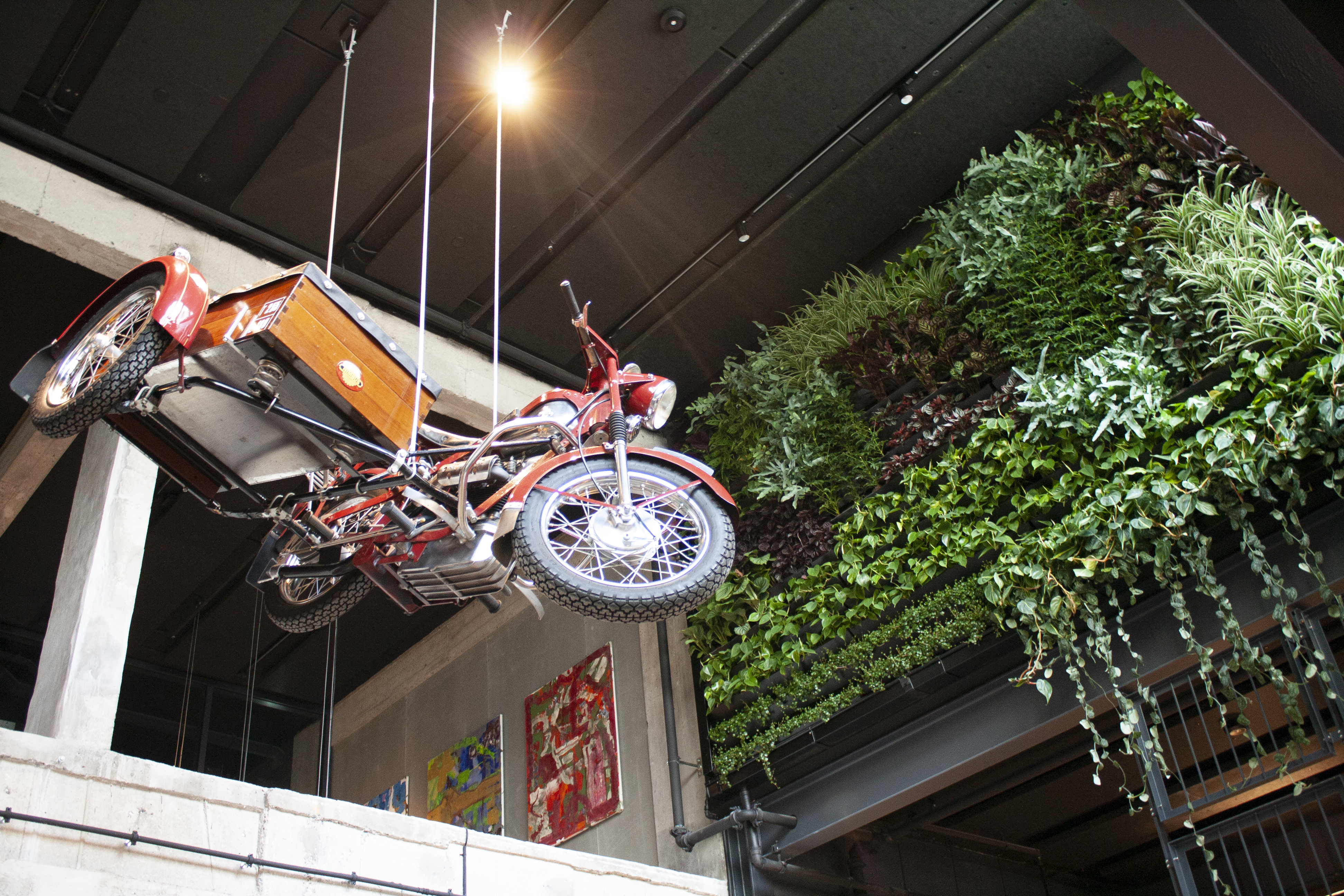 A vintage motorcycle with a sidecar is suspended in a modern interior. To the right, a green plant wall with a mix of green plants covers part of the wall. Strong overhead lighting illuminates the scene, while artworks can be seen on a nearby wall.