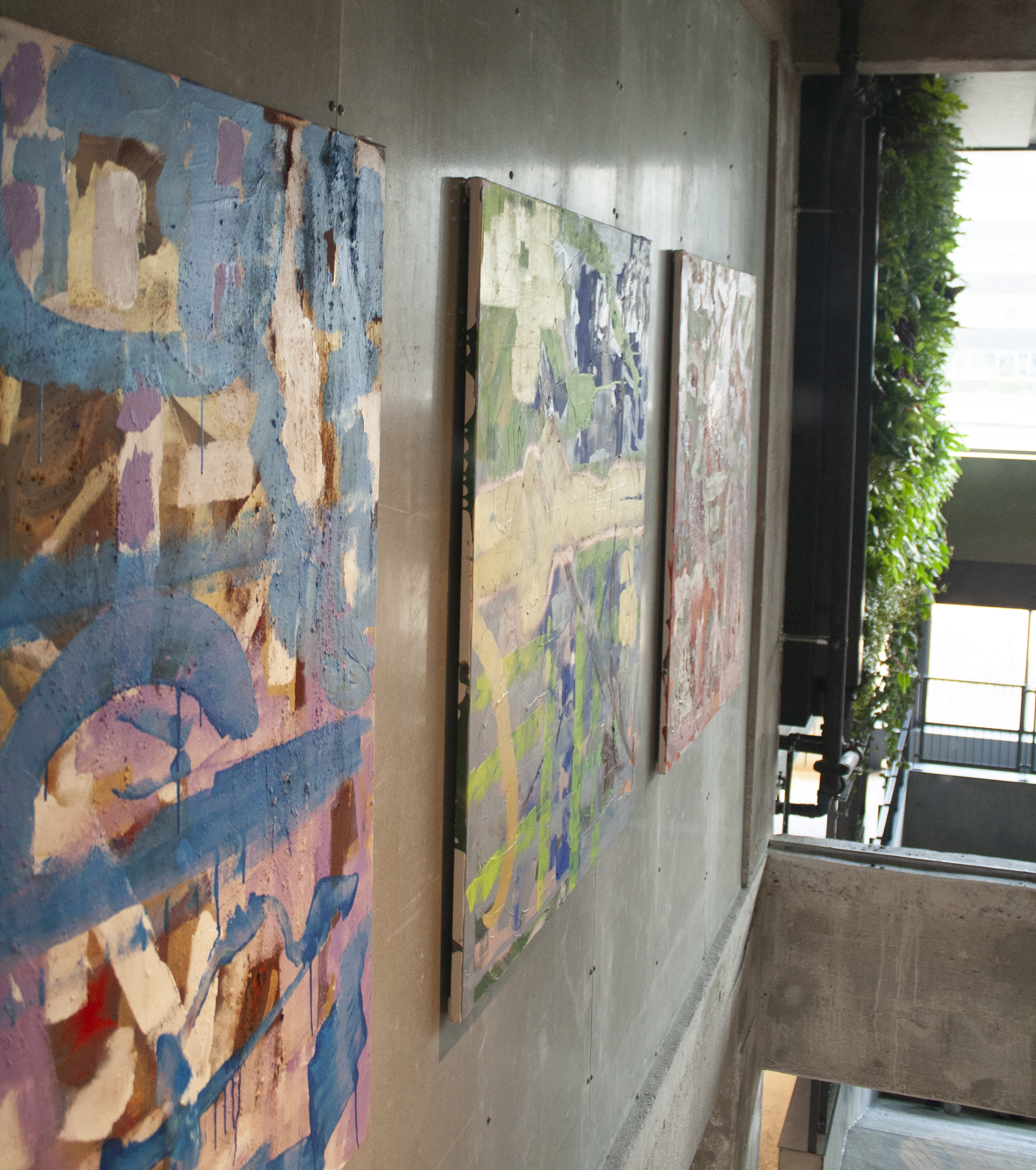 Three abstract paintings with varying colors on a concrete wall. The first is blue-themed, the second has green and yellow hues, and the third features pink and white tones. To the right, the green plants by the window area create a natural contrast to the artistic surroundings.