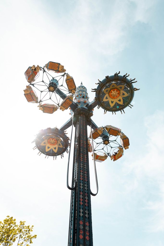 A tall amusement park ride with spinning circular seats ascends into the sky, silhouetted against bright sunlight. Riders are visible with arms raised in excitement. Bright blue sky and light clouds serve as the backdrop.