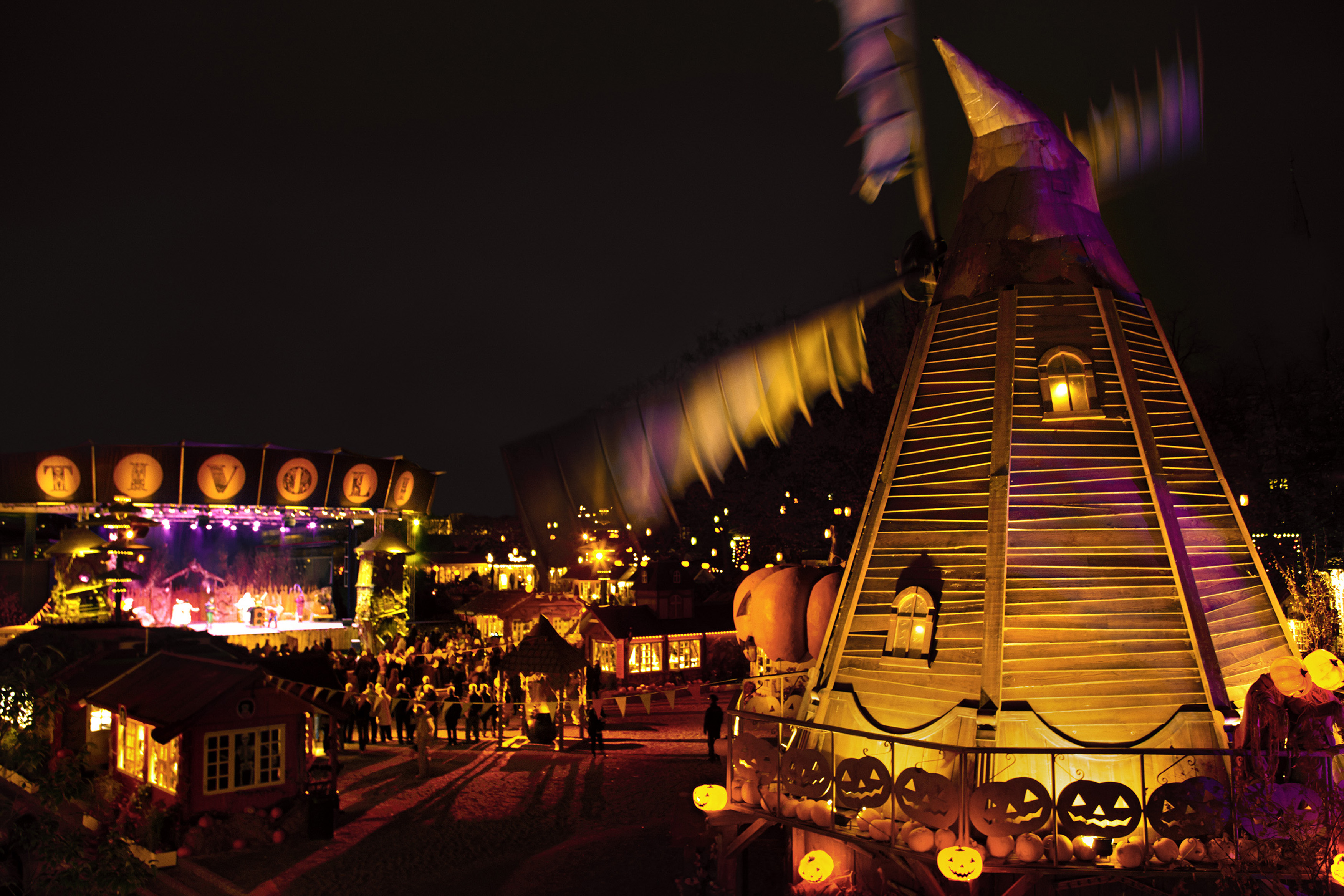 A vibrant nighttime Halloween scene features a lit-up windmill and festive decorations, including pumpkins with carved faces. A stage in the background hosts a performance, and people are gathered around, enjoying the illuminated atmosphere.