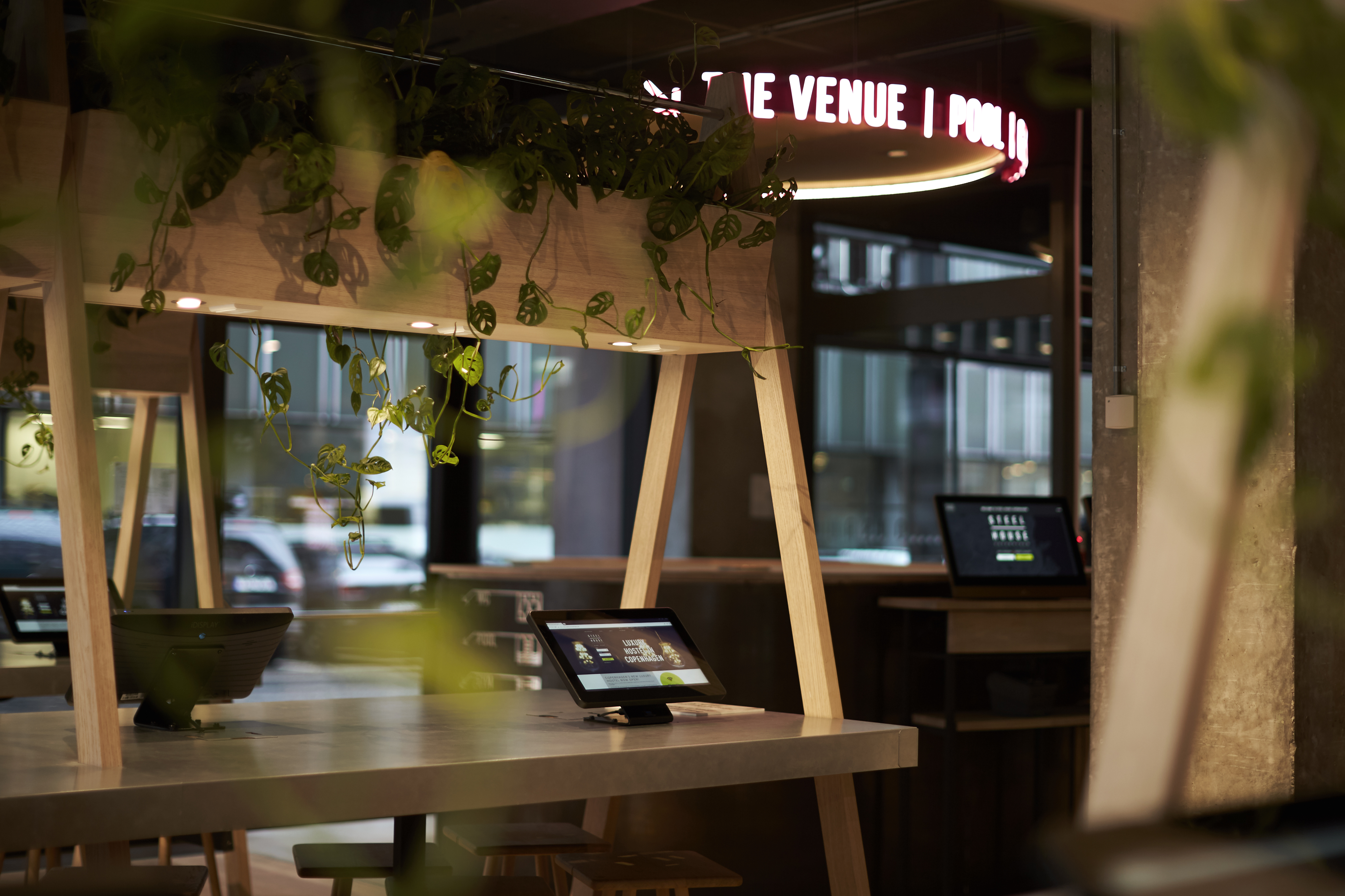 A modern cafe interior featuring wooden tables with built-in screens. Hanging plants add greenery, and a neon sign glows softly in the background. The atmosphere is cozy and tech-friendly.