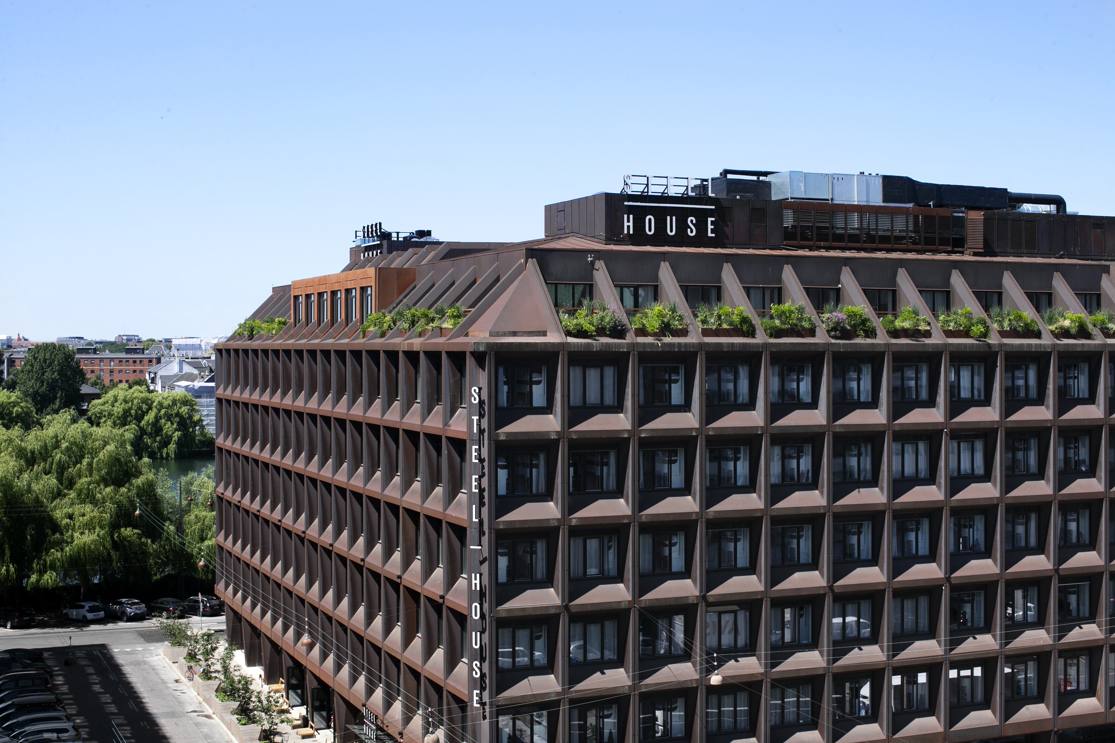 A modern building with a geometric facade and rooftop garden, labeled Steel House. The structure has a grid-like design with greenery on the rooftop. Surrounding trees and a clear blue sky are visible in the background.