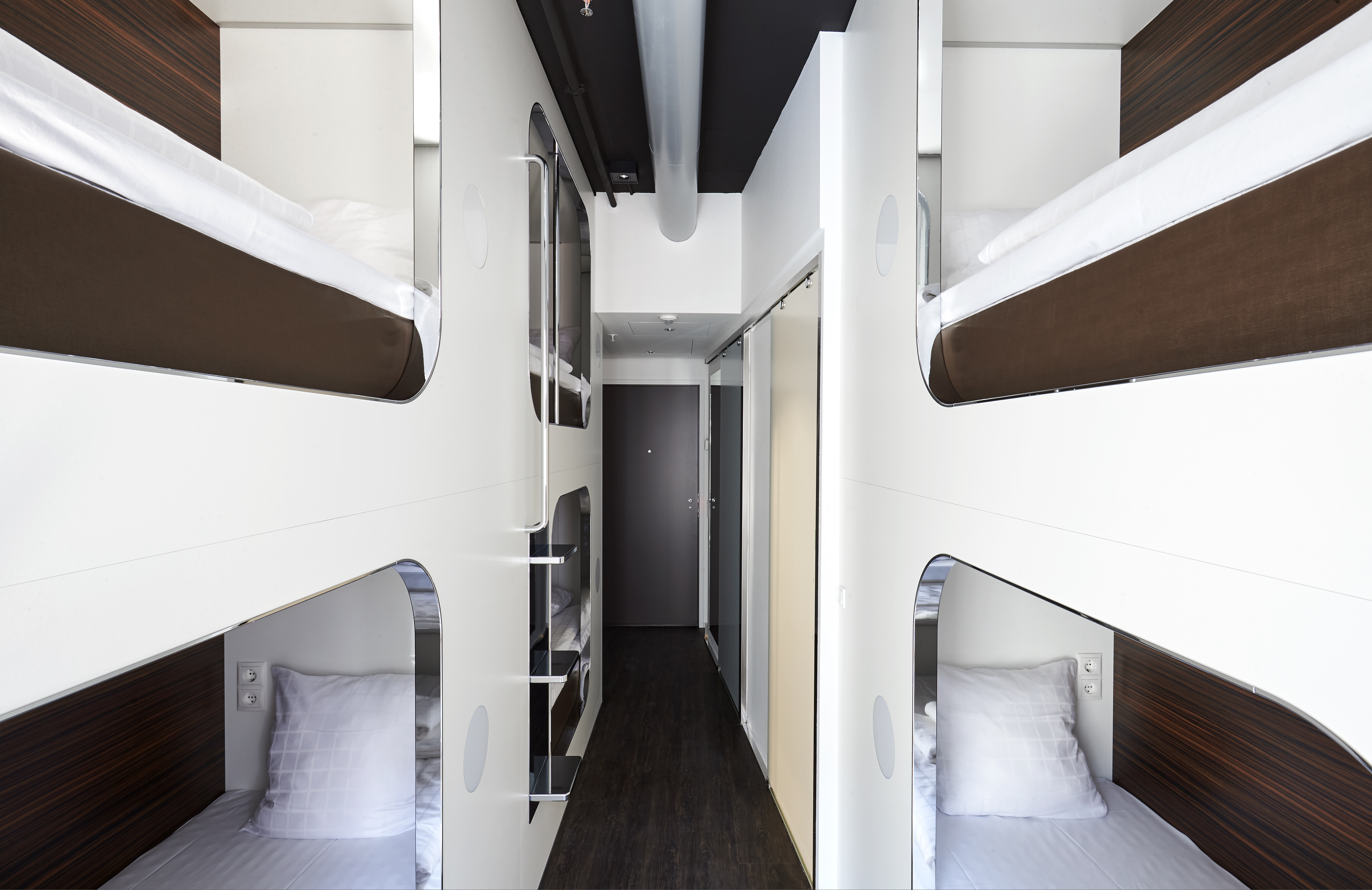 A narrow corridor with modern, minimalist design showcasing four stacked bunk beds on each side. Each bed has wooden accents, white bedding, and a small bedside niche. The space is sleek and utilizes a neutral color palette.