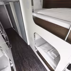 A modern corridor with pod-style bunk beds and sleek white panels on both sides. The wood-textured floor adds warmth to the room, while the recessed lighting highlights the clean, modern design. A closed door is visible at the end of the corridor, and the toilet and bathroom are separate.