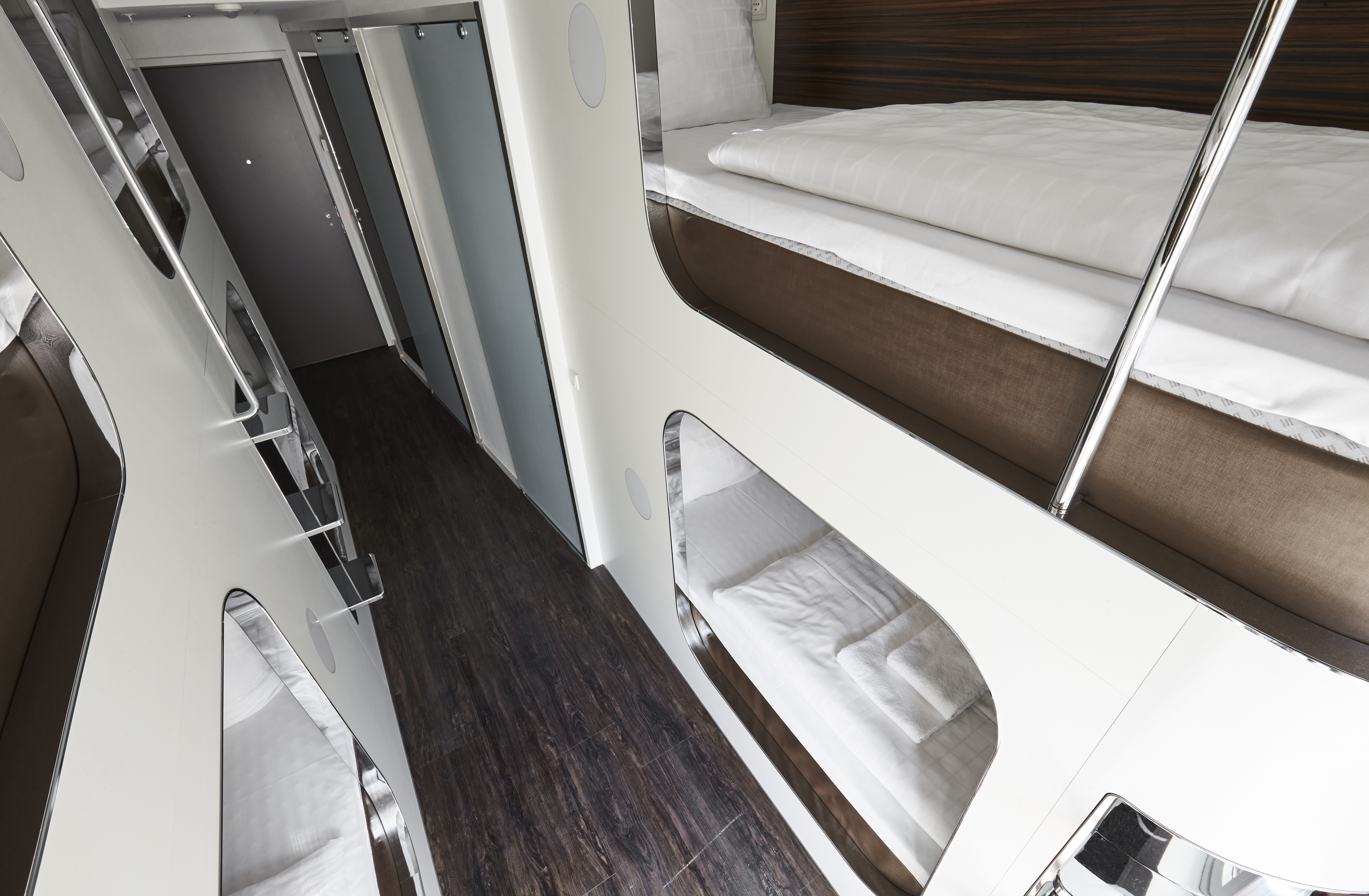 A modern capsule hotel corridor featuring stacked sleeping pods with white bedding on wooden floors. Each pod has a privacy curtain and an individual light. The hallway is clean and minimalistic, leading to a frosted glass door.