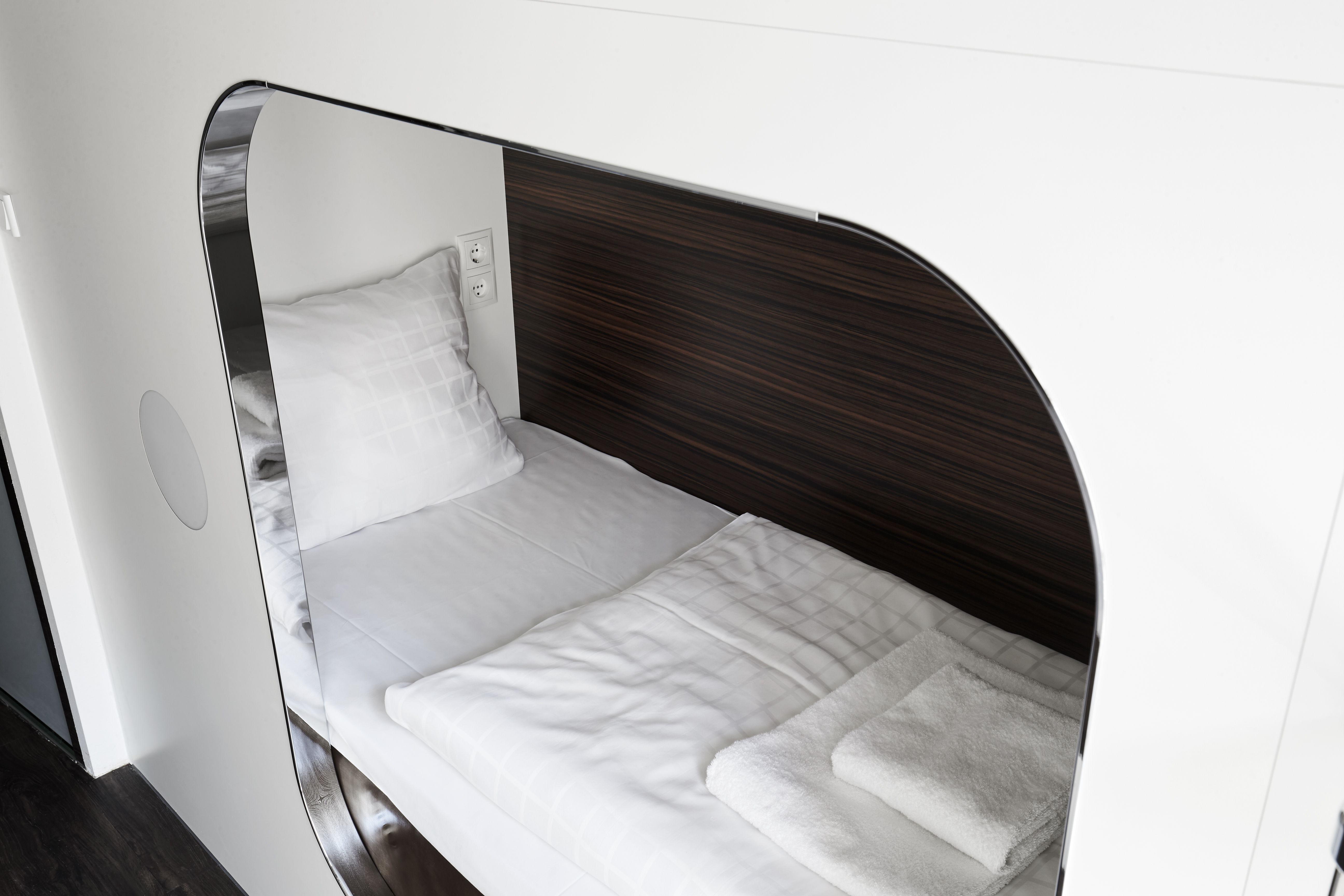 A modern, compact sleeping pod with a neatly made bed, white pillows, and towels in a sleek, wood-paneled space. The pod has a curved, semi-enclosed design with a white frame, offering a minimalist, cozy environment.
