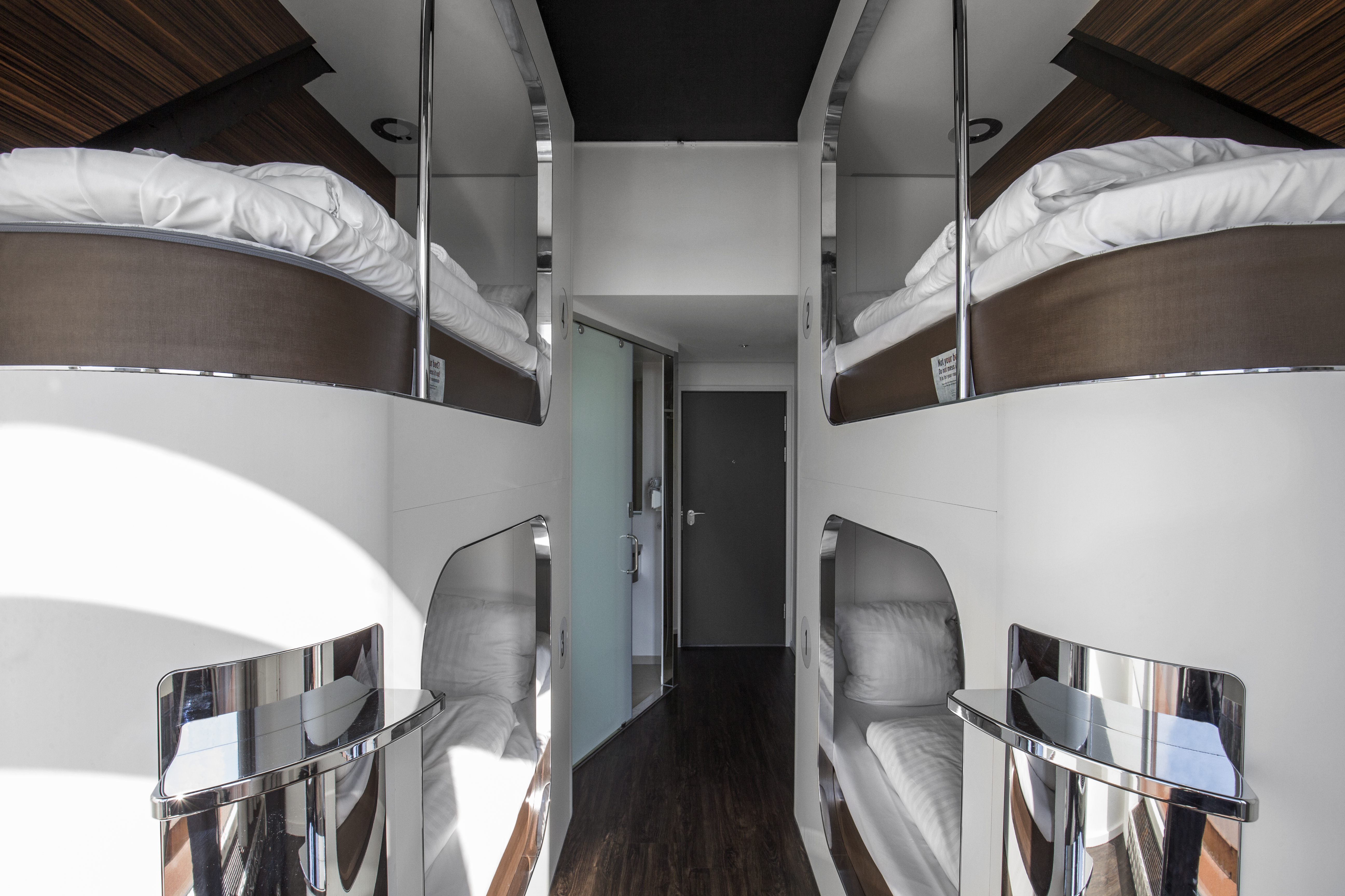Interior of a modern capsule hotel with sleek design. Two mirrored walls on either side feature stacked sleeping pods with white bedding. The dark wooden floor contrasts with the bright, streamlined layout. A door is visible at the end of the hallway.