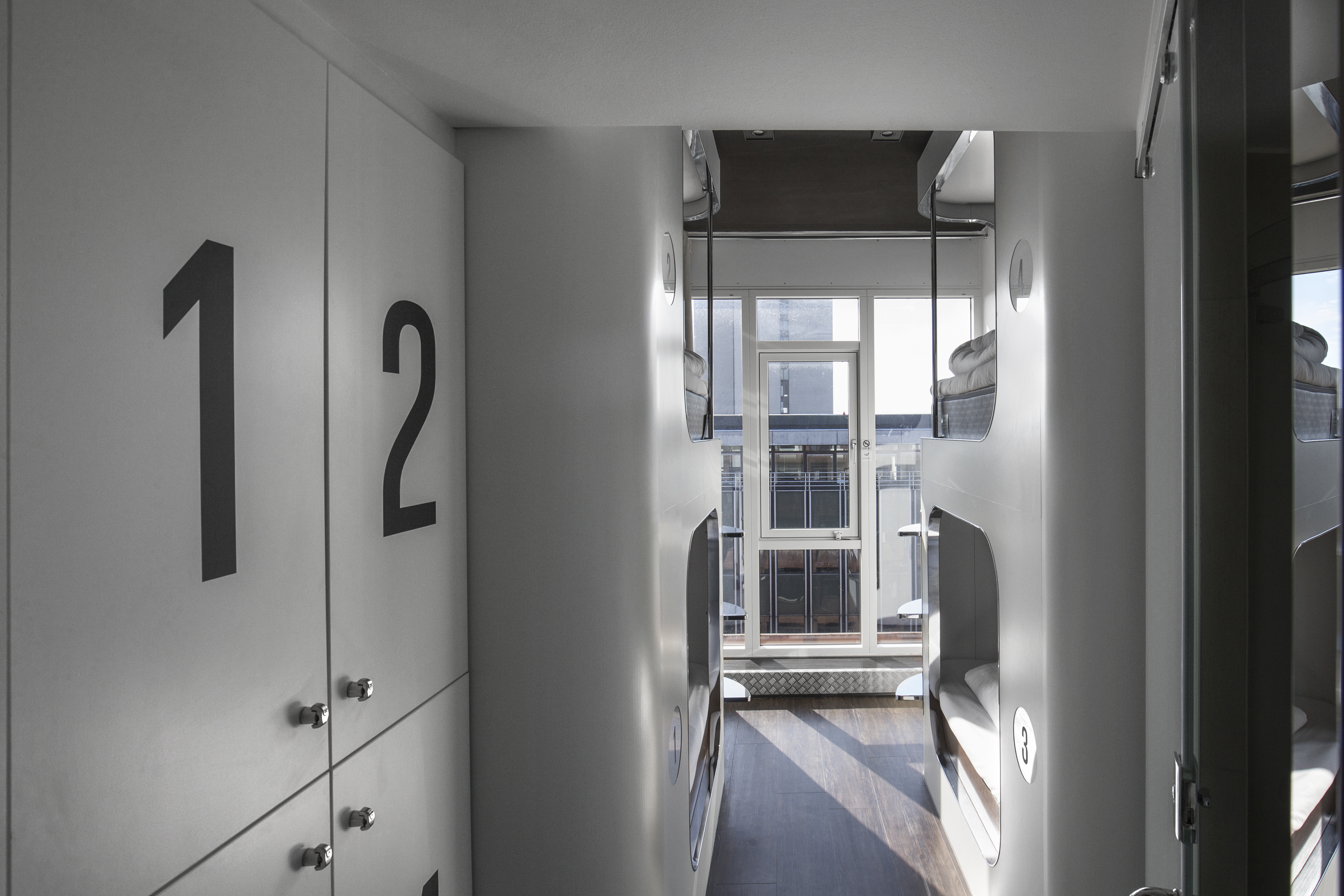 A modern hostel room with bunk beds on either side and numbered lockers, featuring large windows at the end providing natural light. The interior is sleek with a minimalist design.