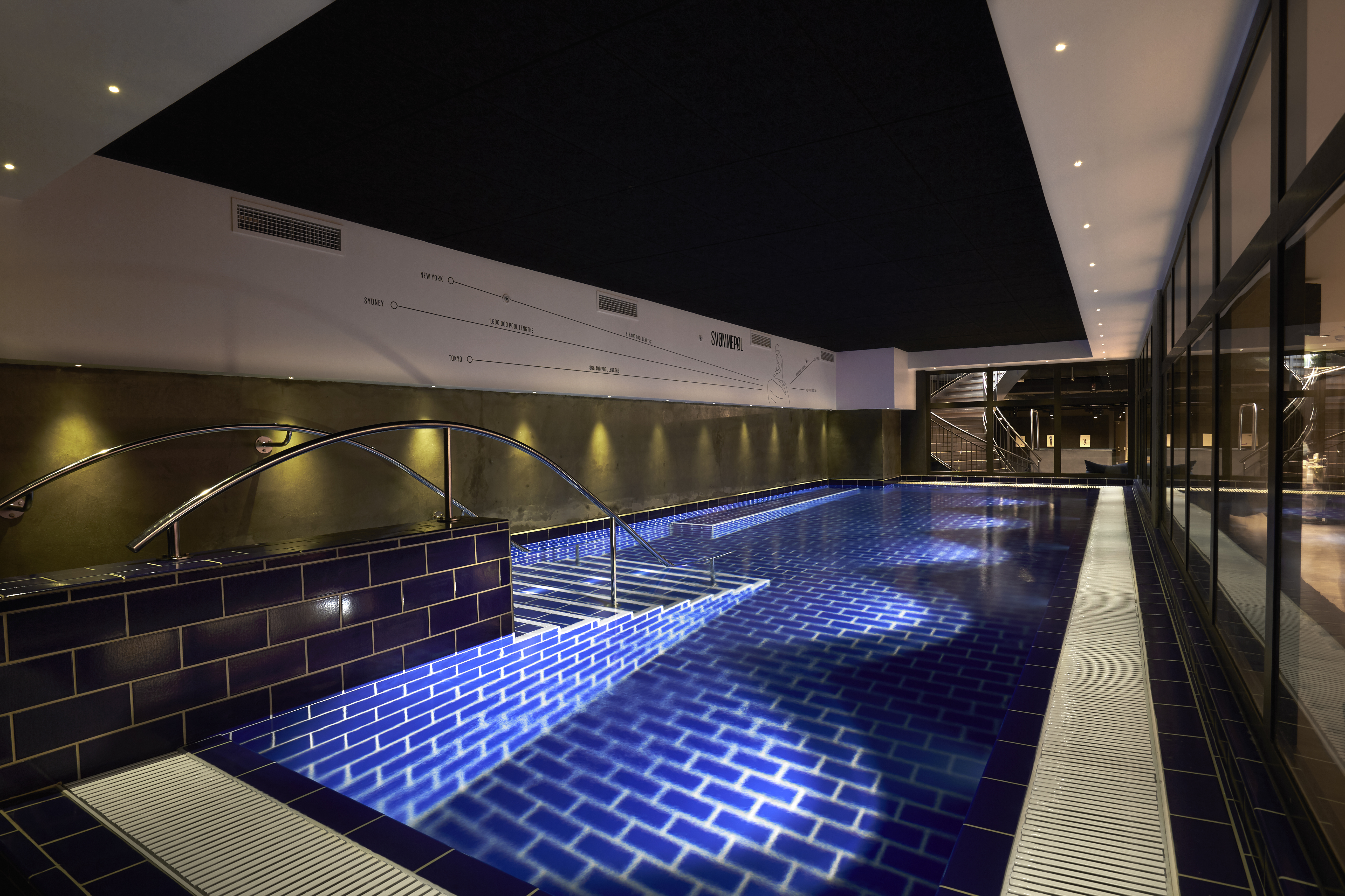 Indoor swimming pool with blue-tiled floor and walls, lit by overhead lights. A metal handrail leads into the pool, and floor-to-ceiling windows on the right allow for natural light. A dark ceiling contrasts with the bright pool area.