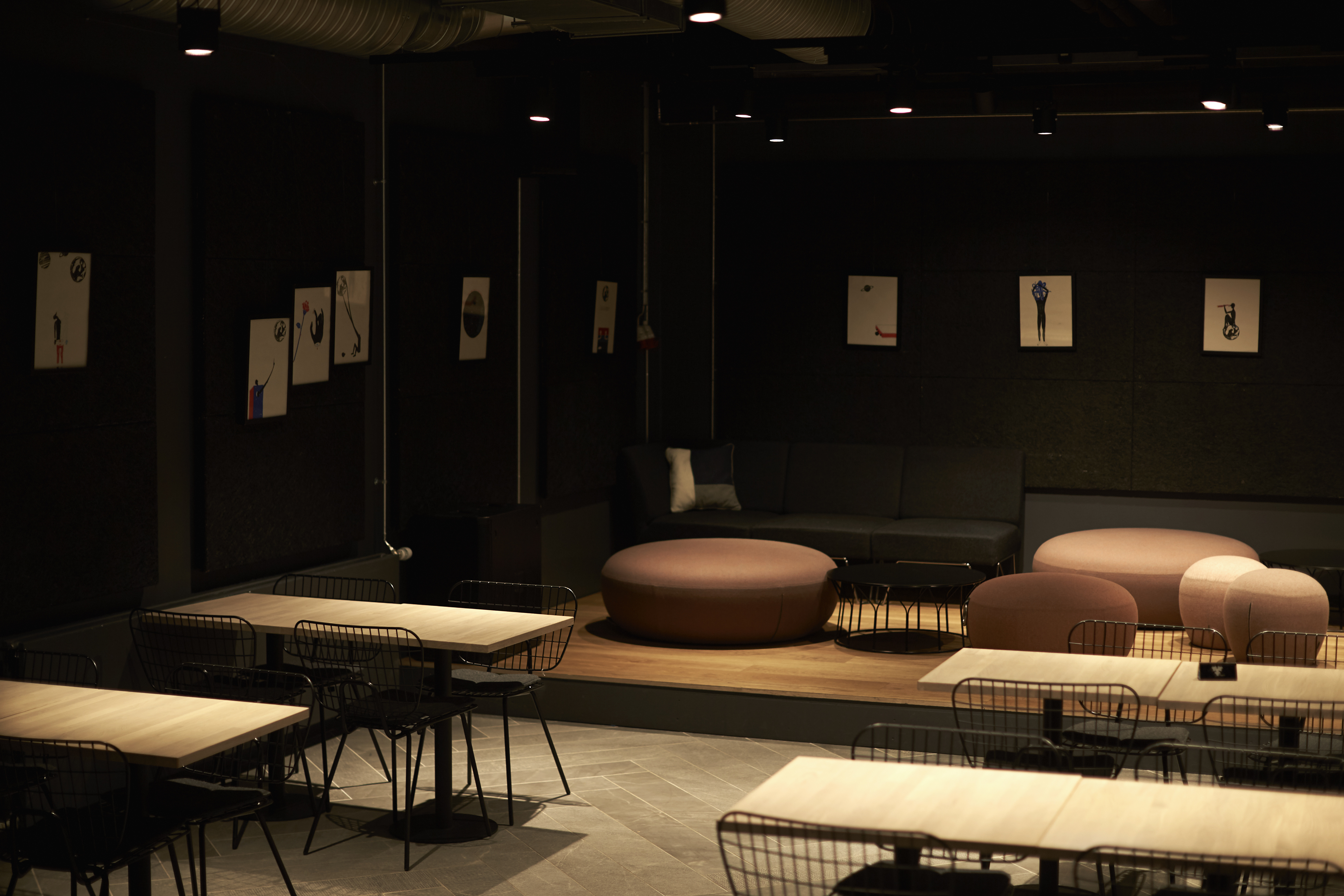 A dimly lit, modern cafe with wooden tables and wire chairs. The space features round cushioned stools and a dark sofa in a corner. Minimalist art pieces hang on the black walls, creating a cozy atmosphere.