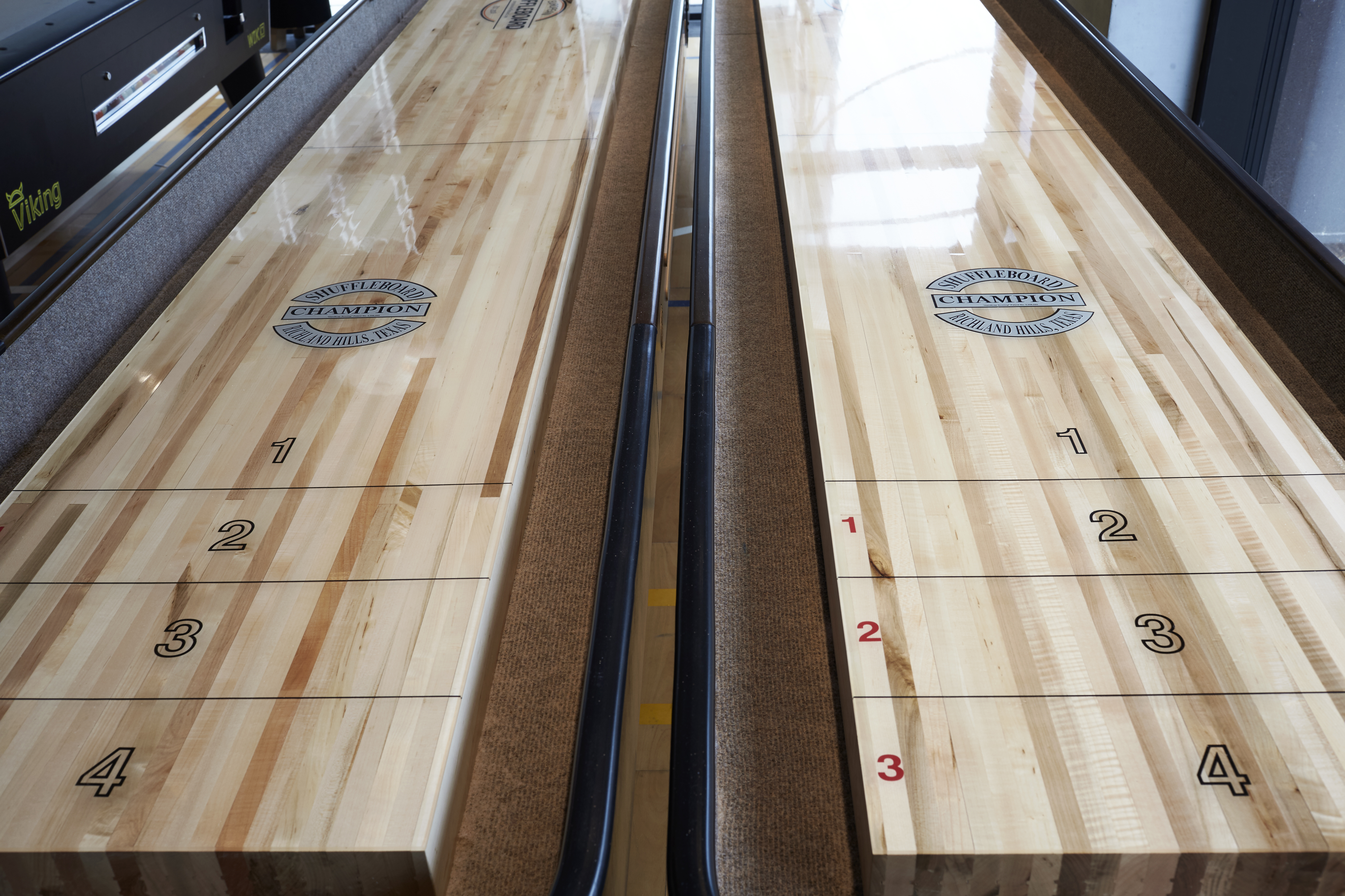 A close-up of two parallel shuffleboard lanes with polished wood surfaces. Each lane has numbered scoring zones marked 1 to 4, with the words Champion Shuffleboard in blue circles on each lane.