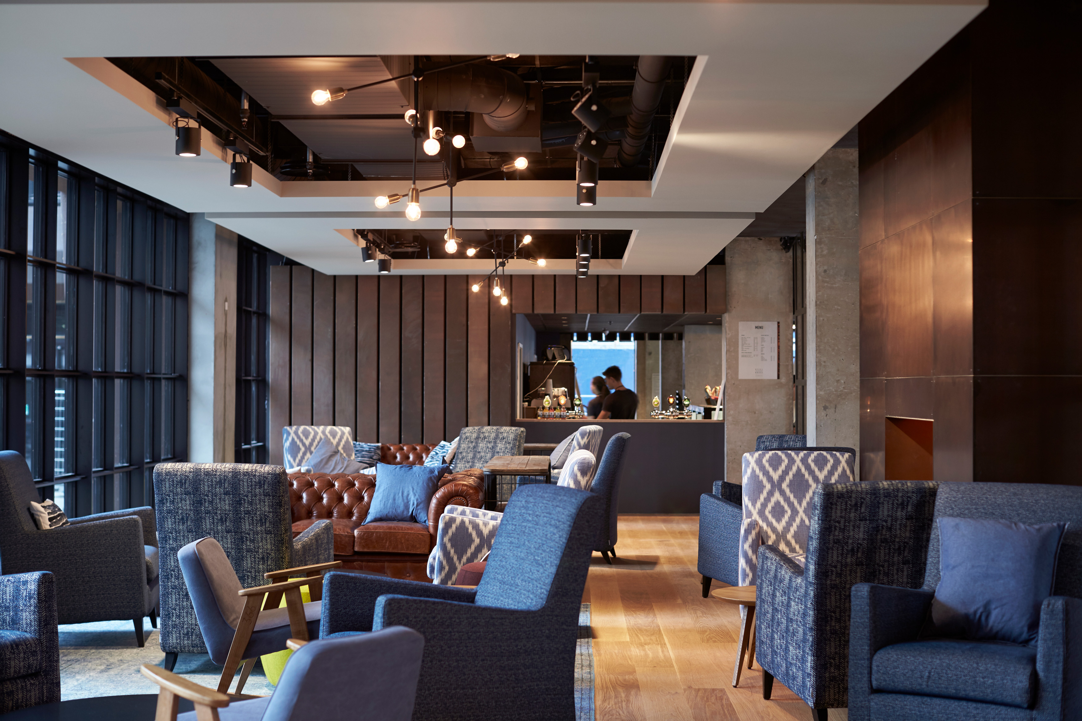 A modern lounge area with blue and brown upholstered chairs, arranged in clusters. The space has wooden floors, large windows, and hanging lights. A person is visible in the background near a counter.
