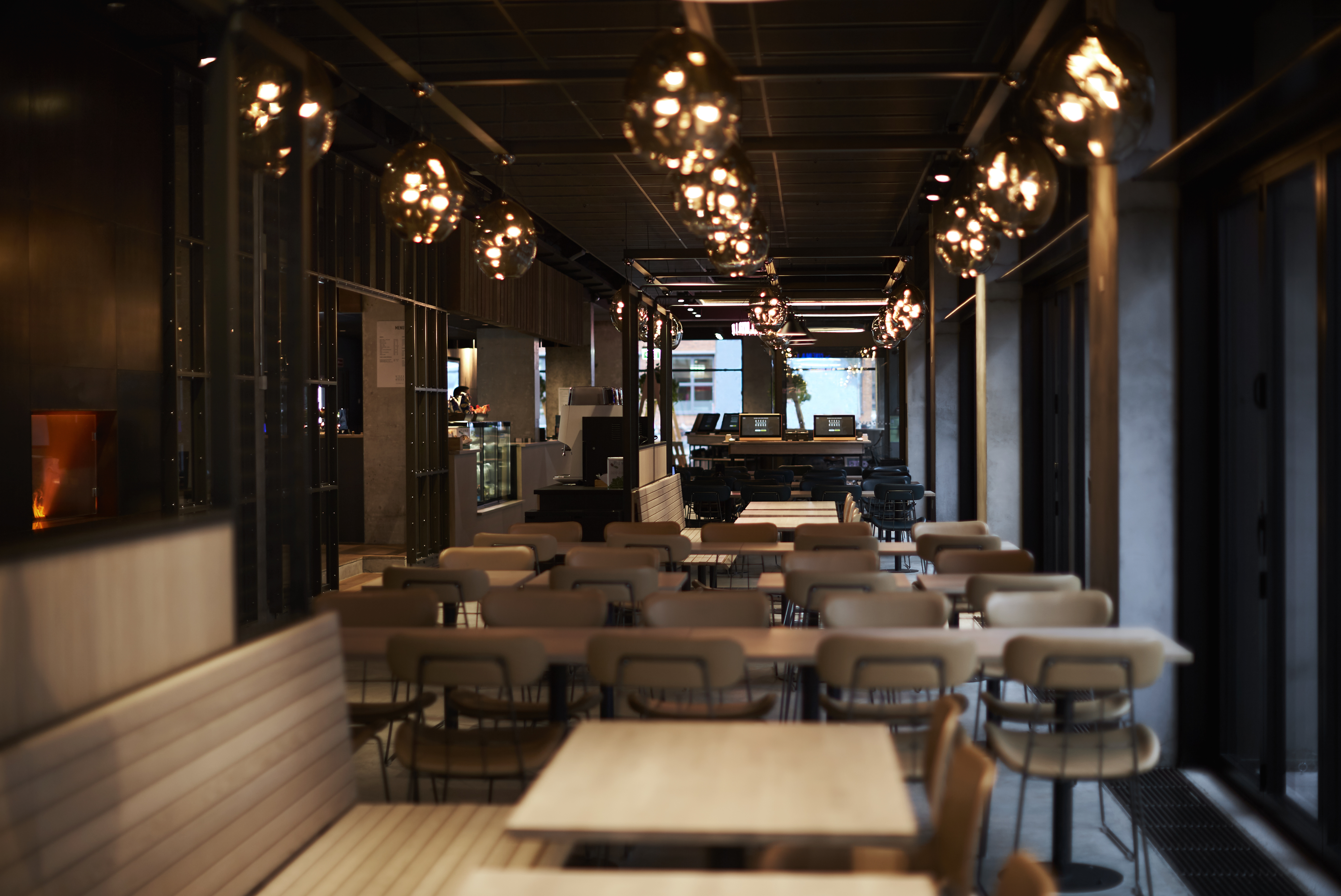 A modern, dimly lit restaurant interior with wooden tables and cushioned chairs arranged in neat rows. Elegant hanging lights create a warm ambiance. The space leads toward a counter area in the background.