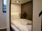 A cosy hostel room with a neatly made bed, white bedding and a folded white towel. The interior features wood panelling and reading lamps. A window offers a view of the city.
