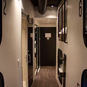 A hallway in the hostel room at Next House with numbered sleeping cabins on both sides. Each pod has a built-in light. A door at the end leads to a toilet and shower cabin. The floor is dark brown, and the walls are white.