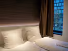 A cosy double bedroom with two white pillows and two duvets on the bed. A window with brown curtains is partially open and shows the city outside. Soft lighting creates a warm atmosphere.