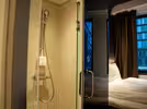 A modern double hostel room with a private shower and chrome shower head on the left. To the right, there is a bed with white bedding near a window with closed curtains. Soft lighting creates a cosy atmosphere.