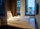 Cosy double hostel room with a neatly made bed with white bedding and towels. A large TV and window offer a view of the city outside. Soft lighting creates a warm atmosphere.