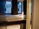 A cosy, modern hostel room with a neatly made bed with white bedding, placed near a large window. Dark curtains frame the window, and the room has a mirror on the side in this stylish space.