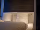 A neatly made bed with white pillows and a duvet is placed against a dark wood-panelled headboard. The headboard has both a power socket and the option to switch the light on and off. Soft lighting highlights the textures, and a window is partially visible to the left, letting in natural light.