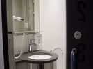 A compact bathroom with a round sink beneath a wall-mounted hairdryer next to a white toilet. The bathroom has beige tiles and a glass shower cabin.