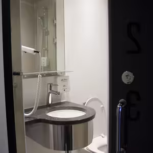 A compact bathroom with a round sink beneath a wall-mounted hairdryer next to a white toilet. The bathroom has beige tiles and a glass shower cabin.