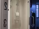 A modern shower cabin with beige tiles, a glass door, a silver-coloured shower head and a soap dispenser on the wall. The shower is located behind a separate door, so the bathroom area is screened off. Numbered cabin doors can be seen to the left, and to the right, a window with a view towards the city is partially visible.