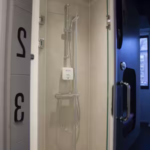 A modern shower cabin with beige tiles, a glass door, a silver-coloured shower head and a soap dispenser on the wall. The shower is located behind a separate door, so the bathroom area is screened off. Numbered cabin doors can be seen to the left, and to the right, a window with a view towards the city is partially visible.
