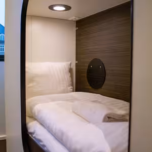 Image of one of the sleeping cabins with a large window in the background. There is a white pillow, towel and duvet on the bed. There is also light inside the cabin.