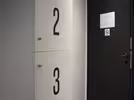 A vertical column with four numbered lockers, labelled 1 to 4, with small metal handles. The lockers are white, spacious and placed next to an open black door. The floor is dark wood, and the ceiling has built-in lighting.