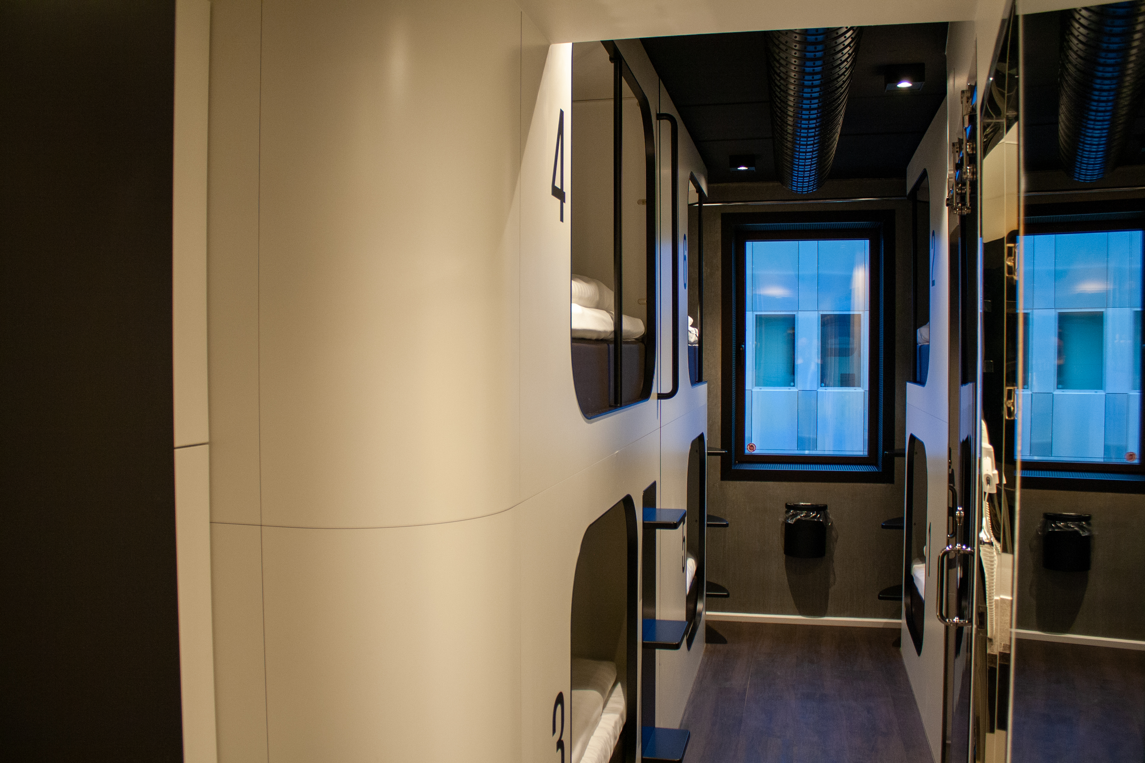 Four beds in a modern hostel room. A window at the end of the corridor lets in natural light. The interior is modern and stylish, with a dark floor and a mirror on the right.