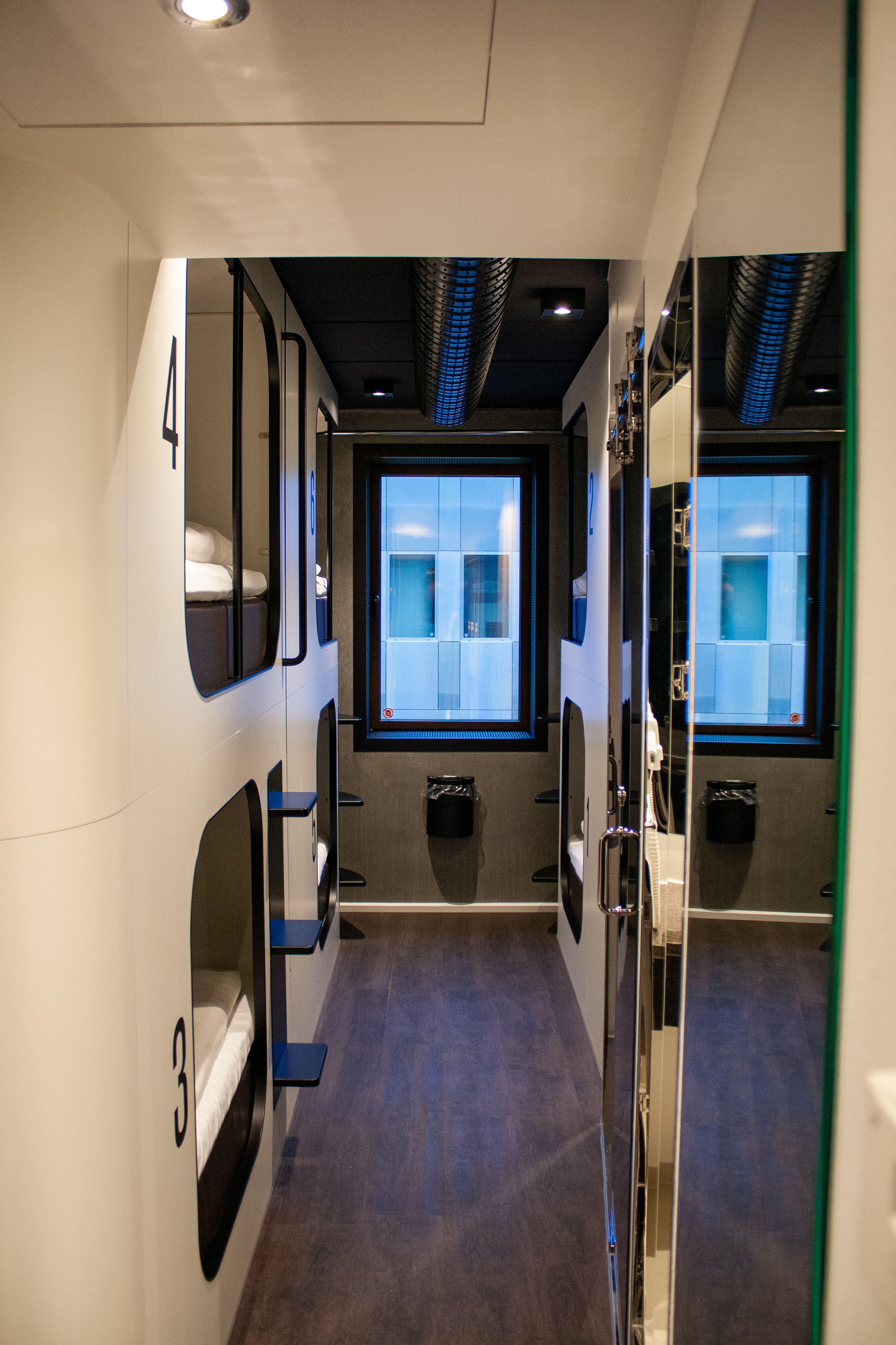 Four beds in a modern hostel room. A window at the end of the corridor lets in natural light. The interior is modern and stylish, with a dark floor and a mirror on the right.