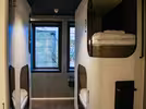 Interior of a modern hostel room with two sets of bunk beds. Each bed has neatly arranged bedding, individual lighting, and a number label. Large windows in the background allow natural light to enter.