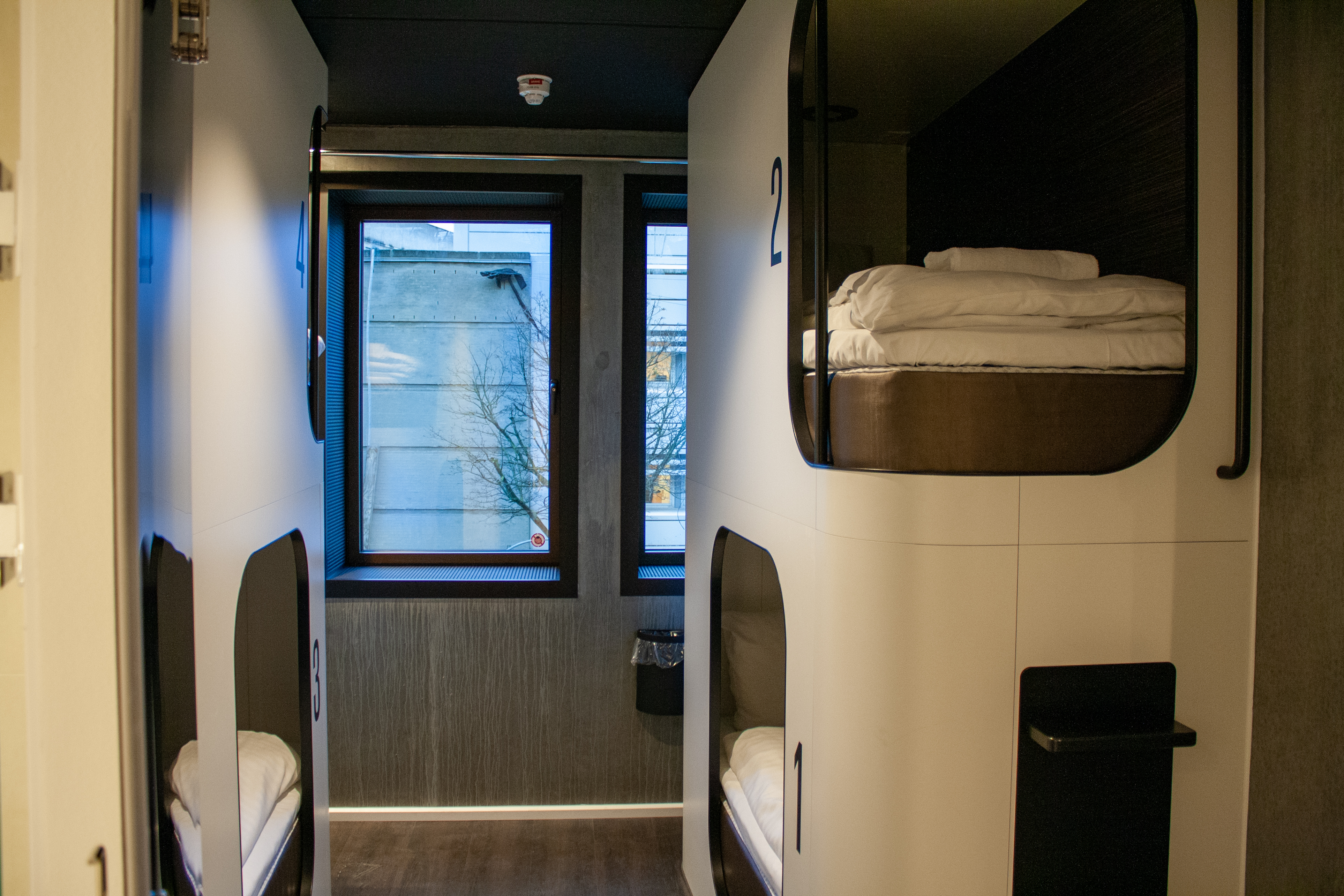 Interior of a modern hostel room with two sets of bunk beds. Each bed has neatly arranged bedding, individual lighting, and a number label. Large windows in the background allow natural light to enter.