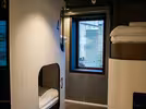 A modern hostel room featuring pod-style bunk beds with numbered compartments and built-in lights. A window provides natural light, showing an urban building view. The room has a sleek, contemporary design with a cozy atmosphere.